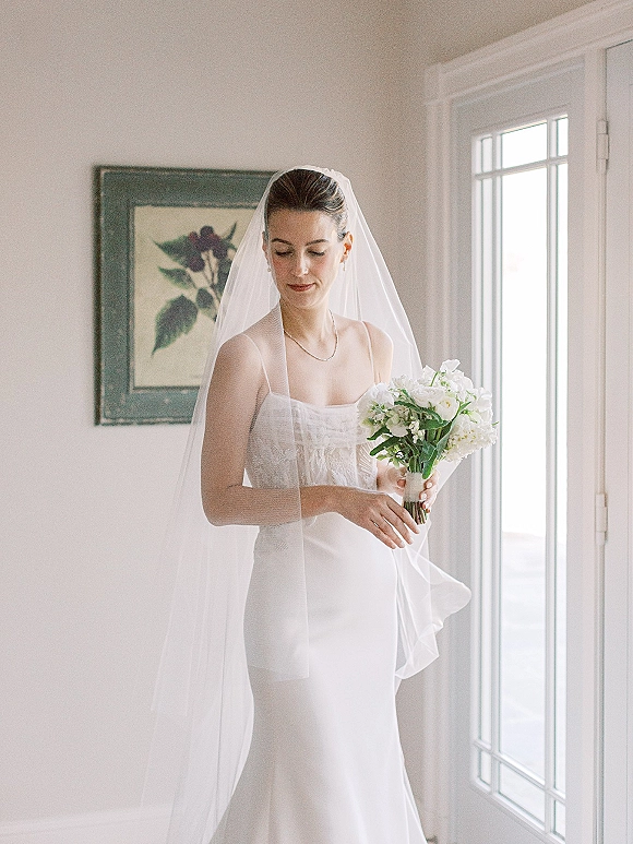 Bridal portrait of a bride holding bouquet of white roses and greenery in a strapless gown and veil, softly lit by a window indoors