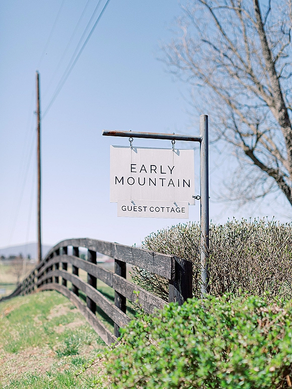 Wedding welcome sign hanging from a wooden post with metal hooks by a rustic fence, set against blue sky, field, and power lines