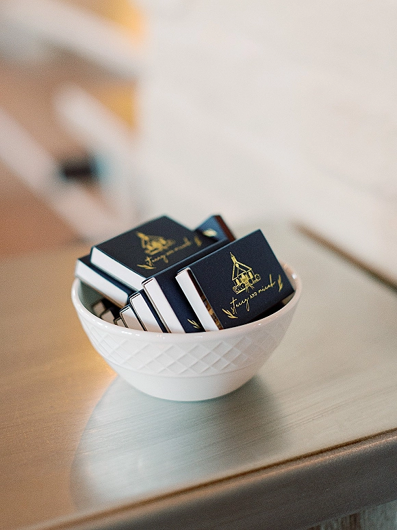 Wedding matchbooks with gold foil print stacked in a ceramic bowl on a tabletop under soft indoor light for reception favors display