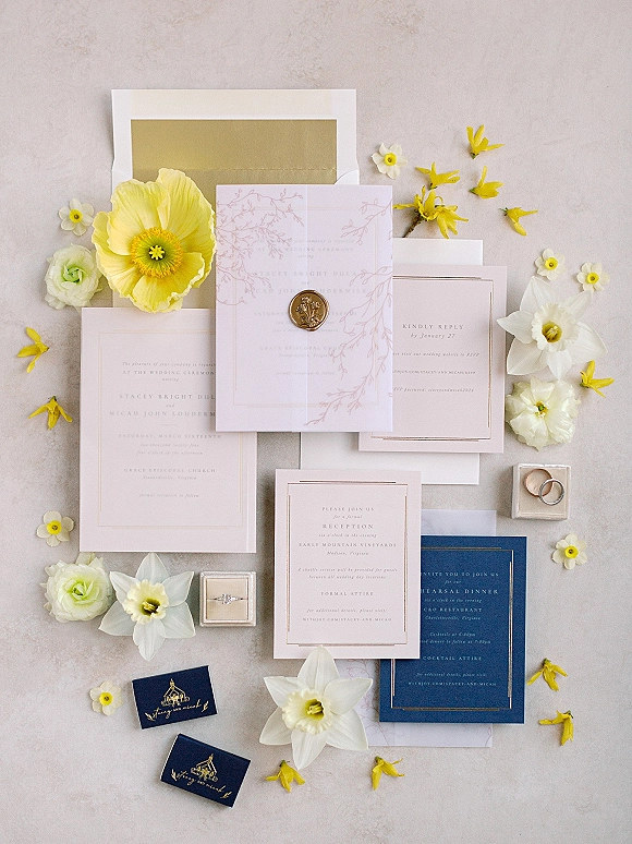 Invitation suite arranged with wax seal envelopes, gold foil details, rings, and flowers on a light stone surface flat lay
