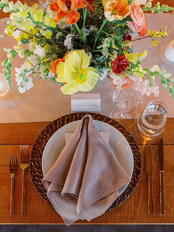 Reception tablescape with wedding place setting, woven charger and folded napkin, plus a colorful floral centerpiece on a wood dining table