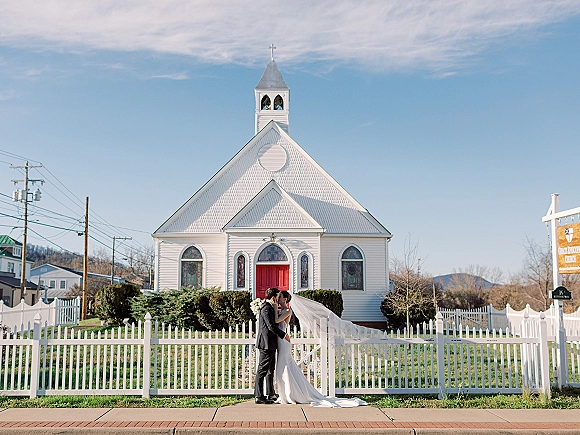 Wedding kiss portrait of bride and groom kissing outside a small church, her veil flowing behind as she holds a bouquet by a white picket fence