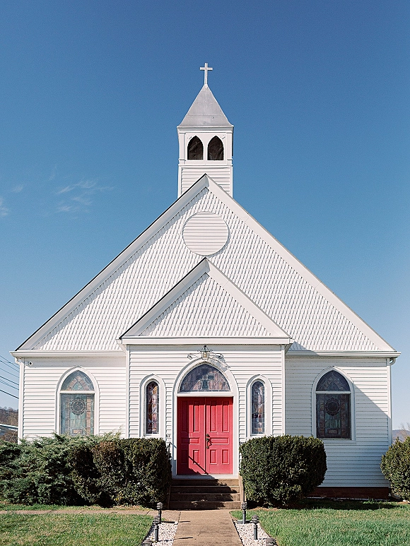 Church exterior with red double doors and stained glass windows on a white steepled facade, lantern lights along the walkway under blue sky