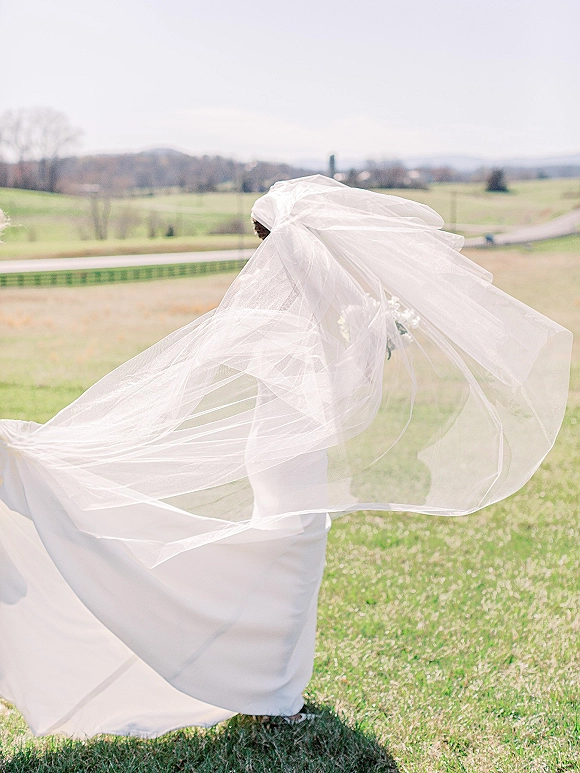 Bridal portrait of a bride in a white wedding dress with a long veil trailing behind, holding a bouquet in a grassy field with rolling hills