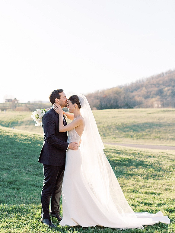 Wedding couple portrait of bride and groom embrace with a forehead kiss, her cathedral veil and long train flowing in a grassy field