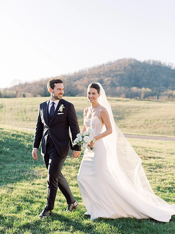 Couple portrait of bride and groom walking hand in hand, her long veil trailing as they smile in a grassy field with hills behind