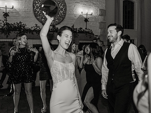 Wedding dance floor scene with bride dancing at the reception as the groom in a vest watches, guests cheering by a brick wall and round mirror