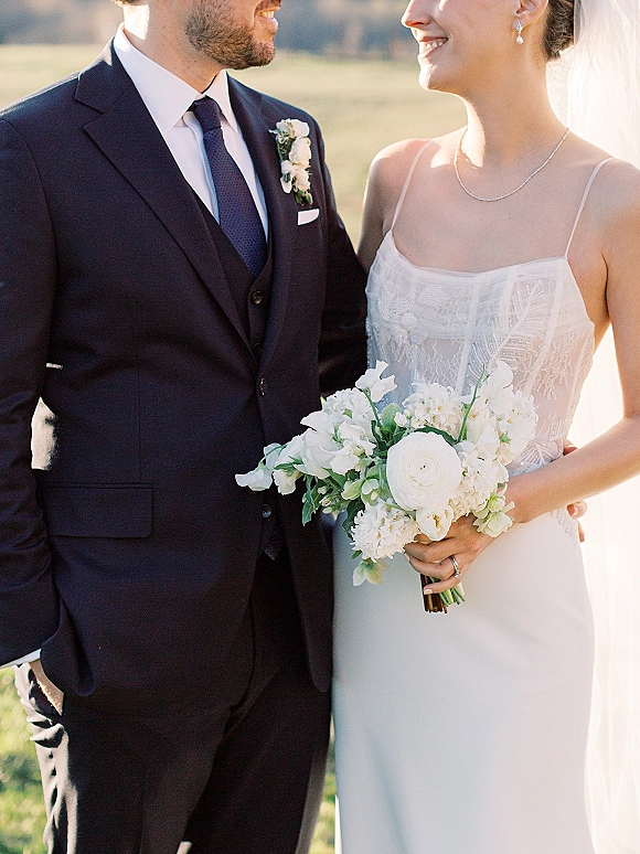 Couple portrait of bride and groom smiling as she holds a white bouquet, her veil draped beside his navy suit in a sunlit field