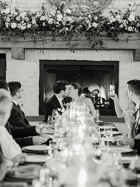Wedding reception kiss as the newlyweds kiss at dinner at a candlelit head table with floral garland, wine glasses, and a brick fireplace backdrop