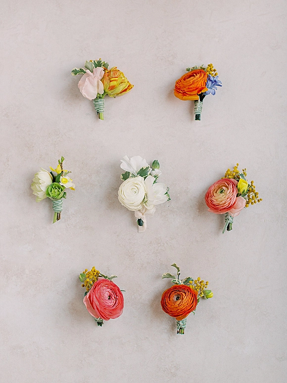 Wedding boutonnieres arranged in a flat lay, featuring ranunculus and greenery with wraps and floral tape on a light stone surface
