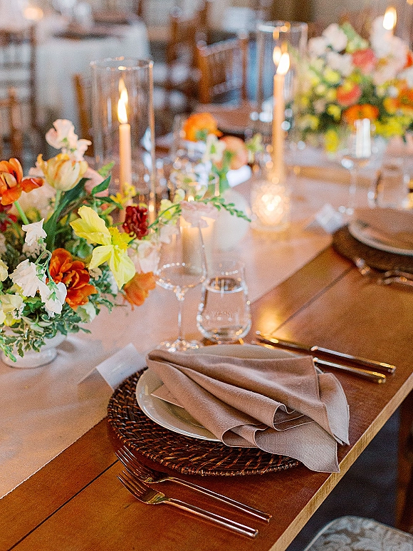 Reception tablescape with wedding table centerpiece of orange-yellow florals, taper candles in hurricane vases, and woven chargers on a wood table