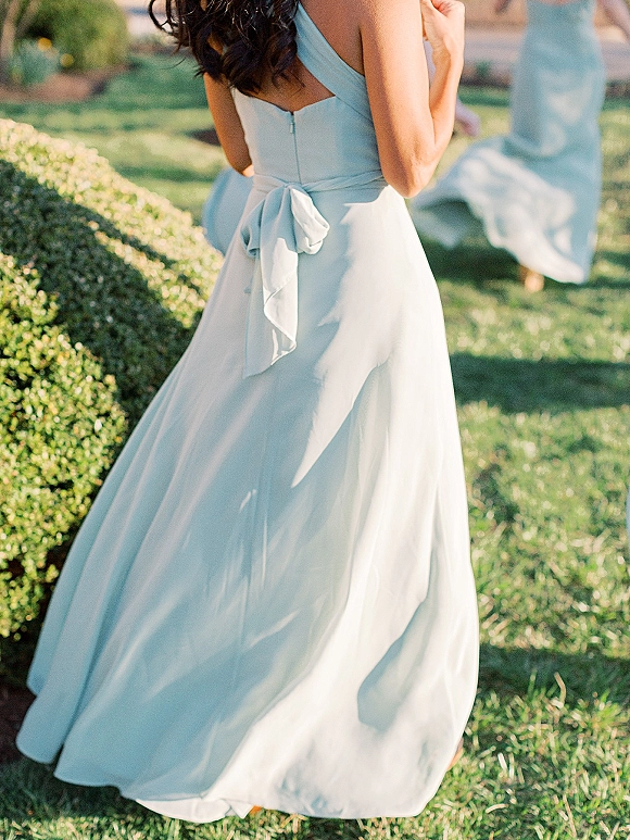 Bridesmaid dress in light blue with a satin bow and zipper back, spaghetti straps and flowing skirt in sunlit garden lawn with hedge