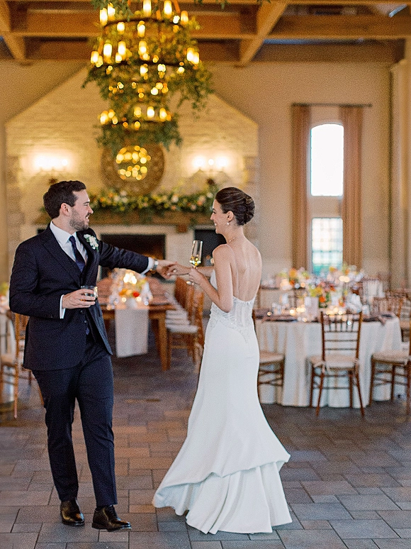 First dance at a wedding reception as bride in a strapless gown and groom hold hands, drinks raised beneath a greenery chandelier by a stone fireplace