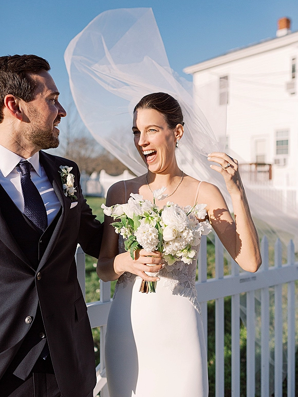 Couple portrait of bride holding bouquet as her veil blows in the wind beside groom in black suit by a white picket fence