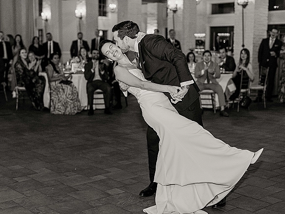 First dance dip as bride in strapless wedding dress and groom in suit kiss on a tiled ballroom floor, guests seated by candlelit tables