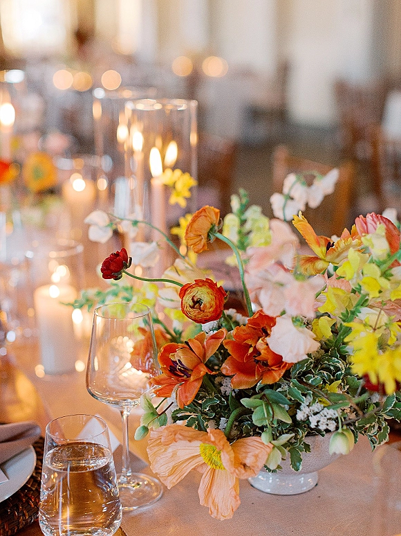 Reception tablescape with wedding table centerpiece of colorful florals, bud vases, taper candles, and woven chargers in warm indoor lighting