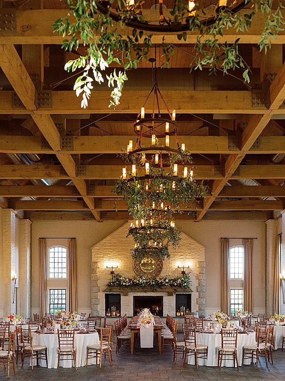 Reception tablescape with long banquet table wedding setup, white linens, wood chiavari chairs, greenery garlands and candles under chandelier by fireplace