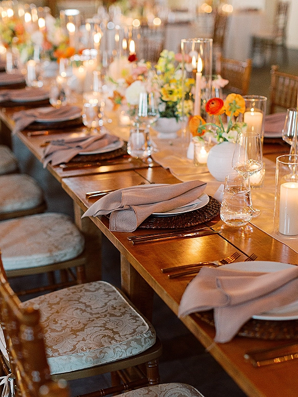 Reception tablescape with wedding dinner table setting, taper candles in glass hurricanes, bud vases, woven chargers on a long wooden table in warm light