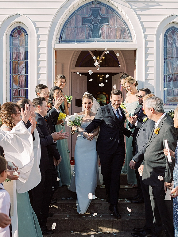 Wedding recessional as bride and groom exit church steps hand in hand, confetti petals falling, veil and bouquet framed by stained glass doorway