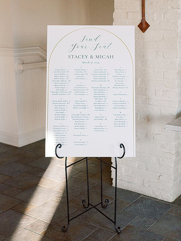 Wedding seating chart with alphabetical seating chart calligraphy on a white poster, framed by a gold arch border on a metal easel by a white brick fireplace