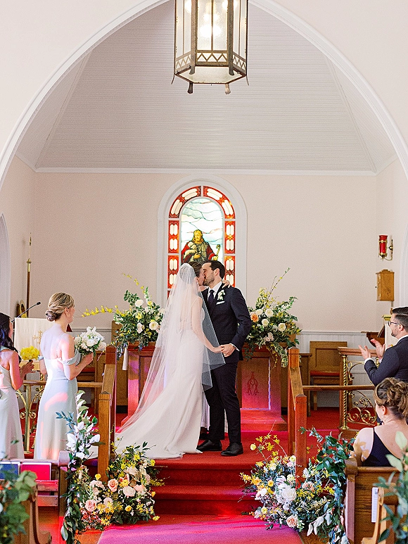 Wedding kiss as bride and groom embrace at the church wedding ceremony altar, long veil flowing beneath stained glass and red aisle runner