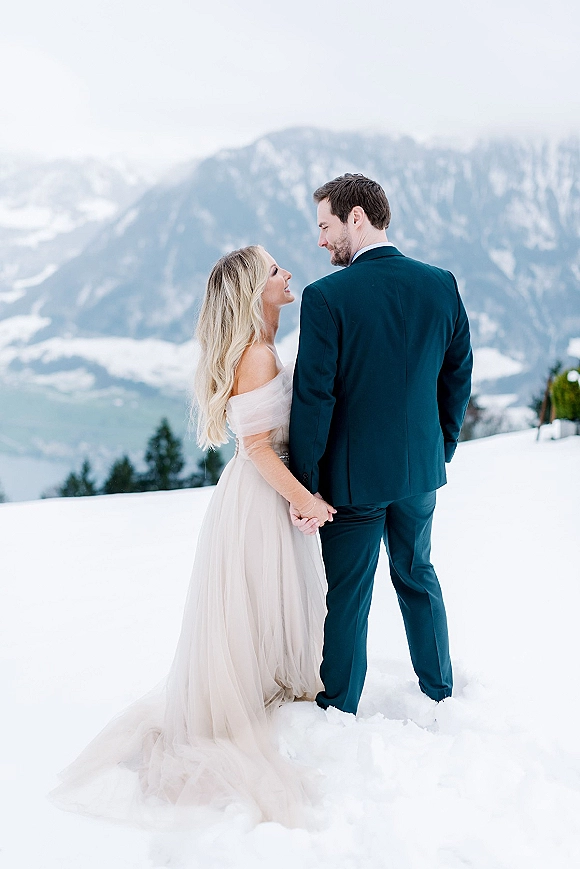 Couple portrait holding hands in a snowy wedding portrait, bride in off-the-shoulder tulle gown beside groom in suit with alpine peaks behind
