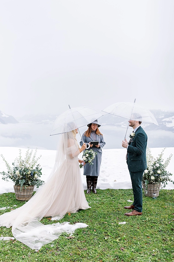 Wedding ceremony with clear umbrellas as bride and groom exchange vows on a snowy mountain lake lawn, veil trailing in overcast light