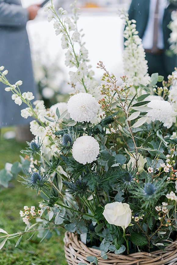 Wedding floral arrangement in a wicker basket with white roses, pompom blooms, blue thistle and eucalyptus on grass, people in suits blurred behind