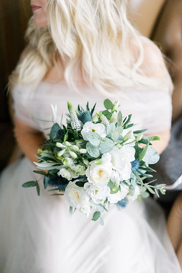 Bridal bouquet of white flowers and greenery with eucalyptus and blue thistle held against a strapless wedding dress in warm blurred interior light