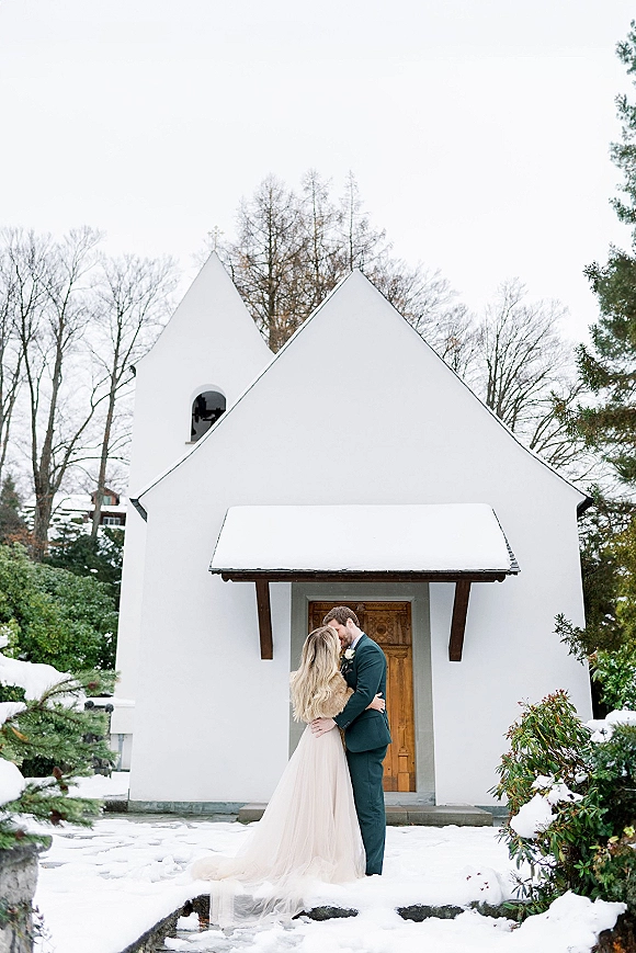 Wedding couple portrait of bride and groom embrace with foreheads touching, bridal gown and boutonniere by a white chapel in snow