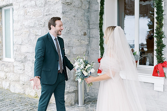 First look moment as bride approaches groom, holding a white-and-green bouquet beside a stone wall and window garland on snow