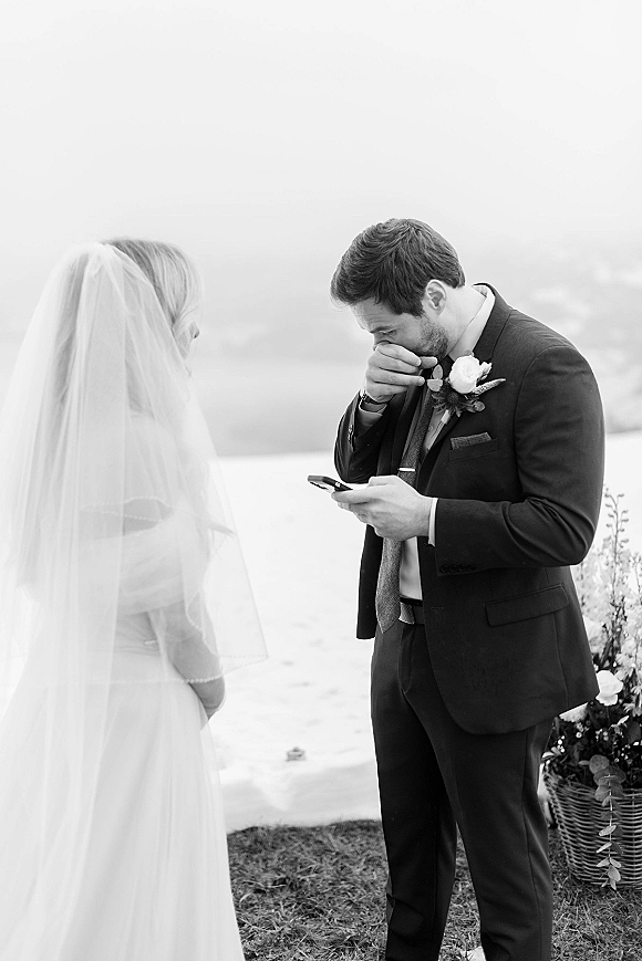 First look moment as bride approaches groom wiping tears while reading vows on his phone by a lakeside shoreline with veil flowing