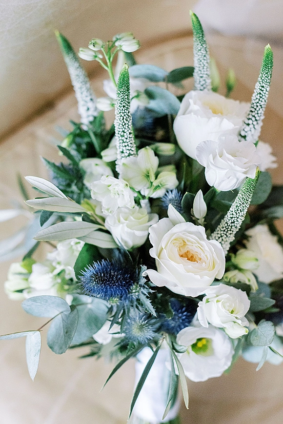 Wedding bouquet of white roses and lisianthus with eucalyptus and olive leaves, blue thistle accents, resting on a wooden surface