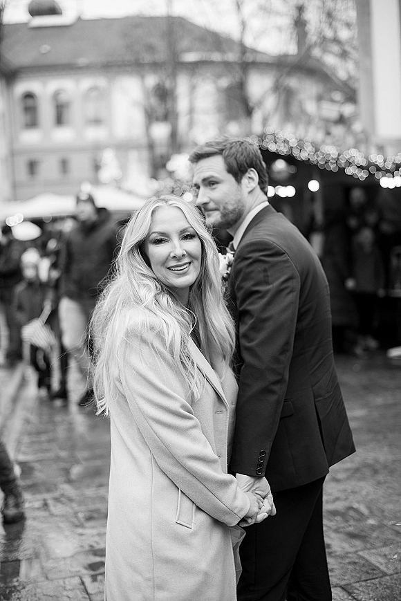 Couple portrait in a black and white wedding portrait holding hands on a city street under string lights, groom in dark suit with boutonniere