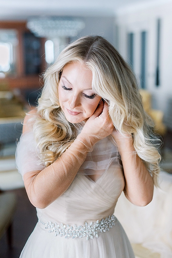 Bridal portrait of a bride getting ready, putting on earrings in a strapless wedding dress with a beaded belt in window light indoors