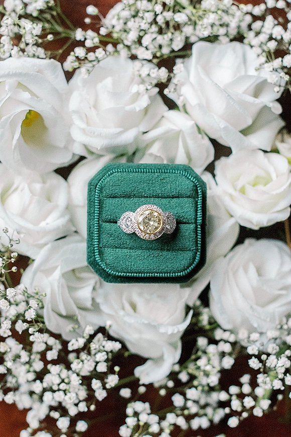 Engagement ring in a velvet ring box, halo diamond with side stones nestled in white roses and baby's breath on a wood surface