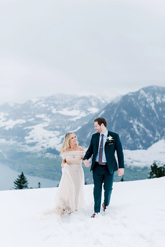 Couple portrait of bride in off-the-shoulder gown and groom in suit holding hands in snow, with mountains, lake, and evergreens behind