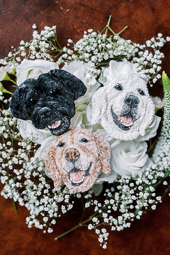 Pet boutonniere with embroidered dog face pins nestled among white roses, baby's breath, and greenery on a brown surface