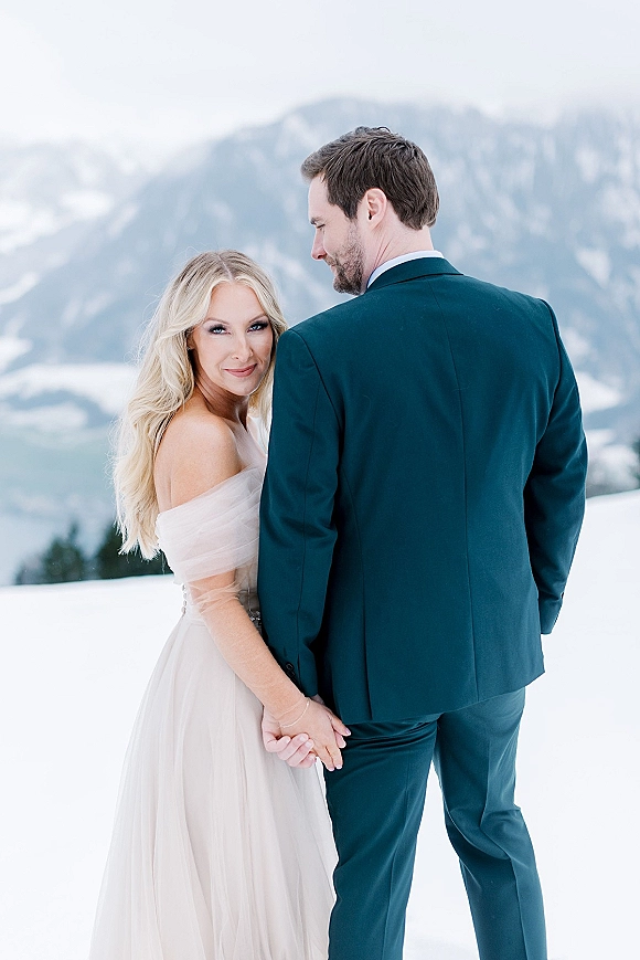 Couple portrait in a snowy wedding portrait, bride in off the shoulder tulle dress holding groom’s hand with mountains and evergreens behind