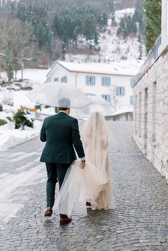 Wedding couple portrait of bride and groom walking away hand in hand under clear umbrellas on a snowy cobblestone street by stone buildings