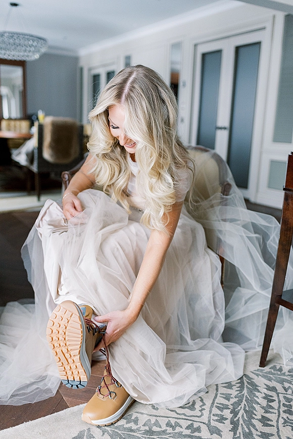 Bridal getting ready bride putting on shoes, sitting in a chair lacing hiking boots beneath a tulle wedding dress in a cozy room