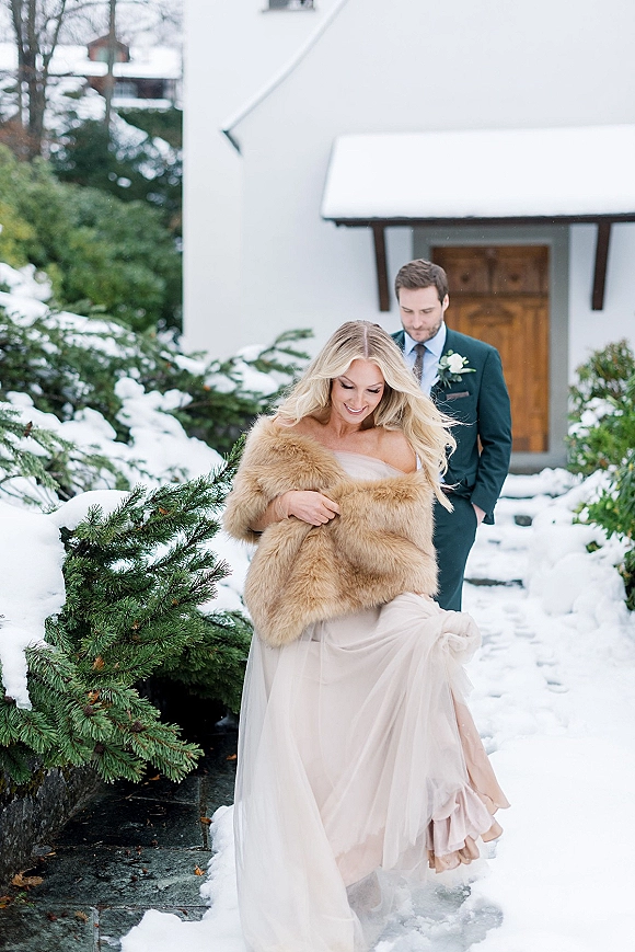Couple portrait of bride in a strapless wedding dress and fur wrap with groom in suit, walking on a snowy stone path by evergreens