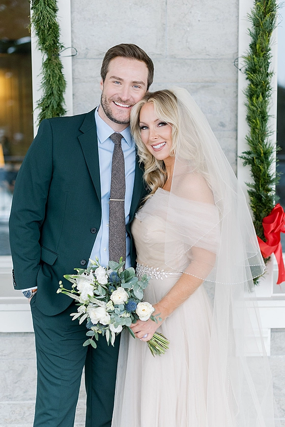 Couple portrait of bride and groom smiling, her veil and bouquet framed by evergreen garland with red ribbon at a stone doorway