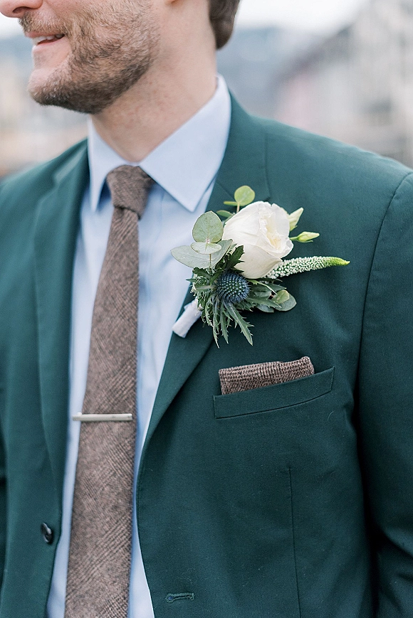 Groom boutonniere with a white rose and blue thistle pinned to a green suit jacket beside a pocket square, outdoors blurred background