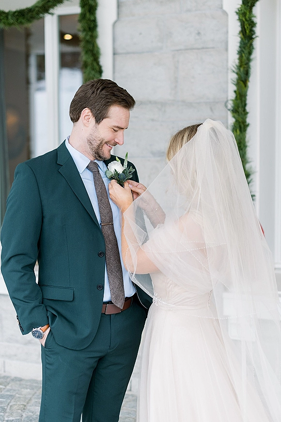 Wedding first look as bride adjusting boutonniere on groom’s navy suit, her veil draped beside stone column and greenery garland doorway