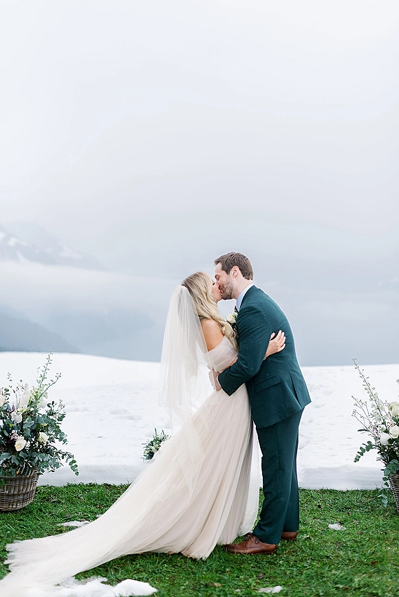 Wedding kiss portrait of bride and groom kissing, her veil trailing over dress train beside floral baskets, with snowy lake and mountains behind