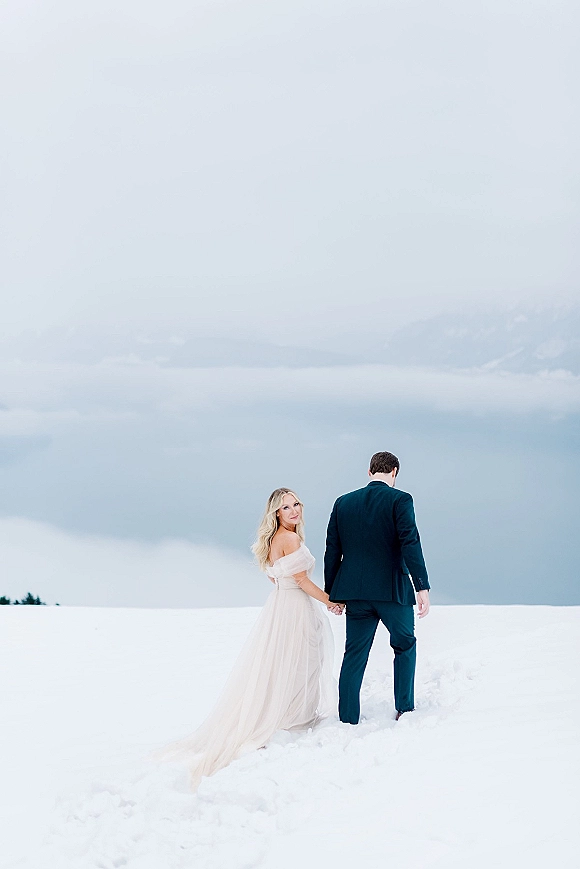 Couple portrait in snowy wedding photos, bride in off-the-shoulder gown and groom in tuxedo holding hands in a mountain snowfield