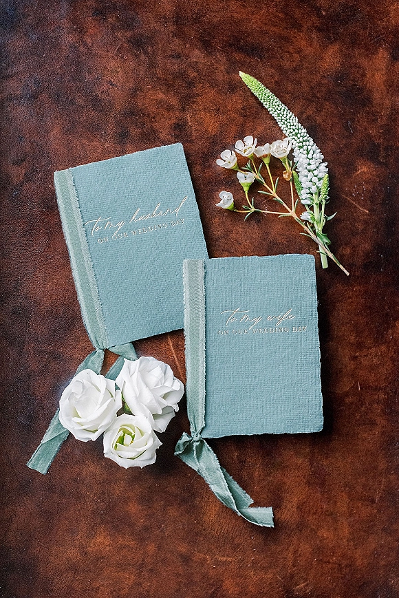 Wedding vows books with linen covers and gold foil lettering, tied with velvet ribbon beside white roses on a brown leather surface