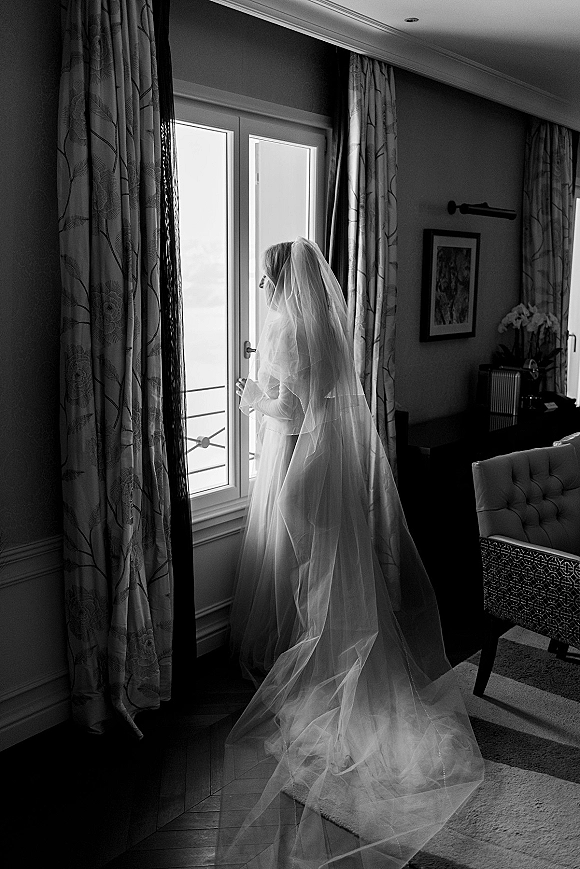 Bridal portrait of a bride looking out window in a bridal gown, her long wedding veil trailing behind in a softly lit room