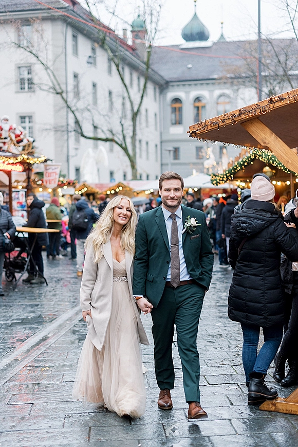 Couple portrait of bride and groom walking hand in hand, her wedding dress under a long coat, at a wet cobblestone market with string lights